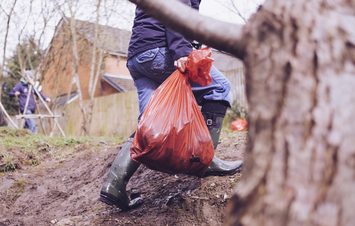 Volunteer in wellingtons carries a litter orange sack and walks behind tree