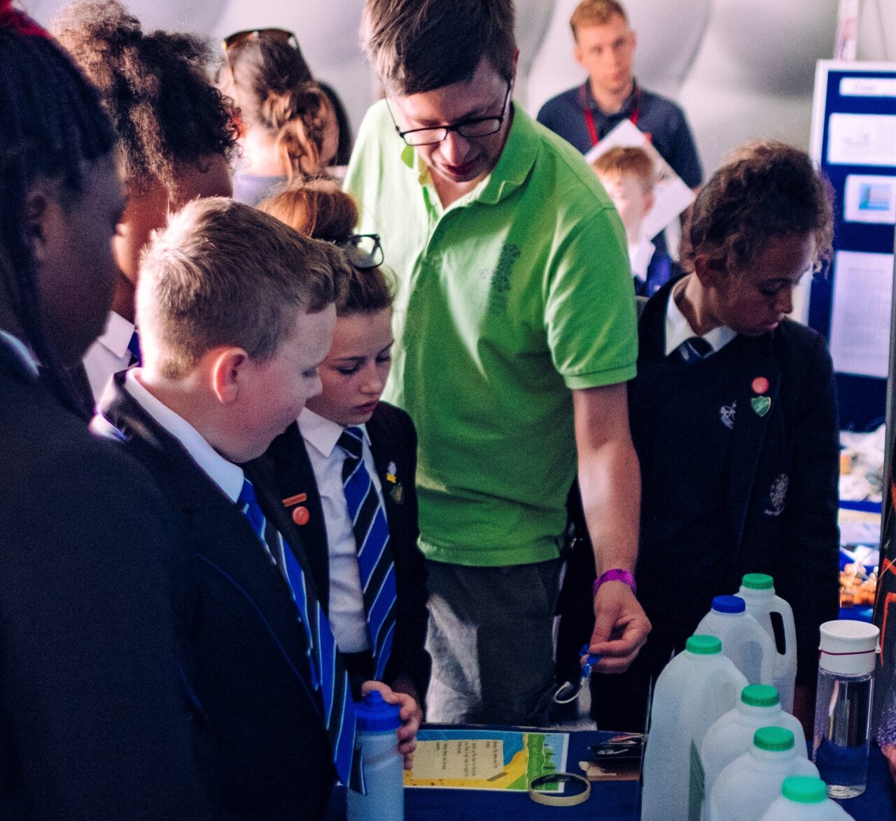 A person wearing a green polo shirt is pointing at empty milk bottles with three young learners watching.