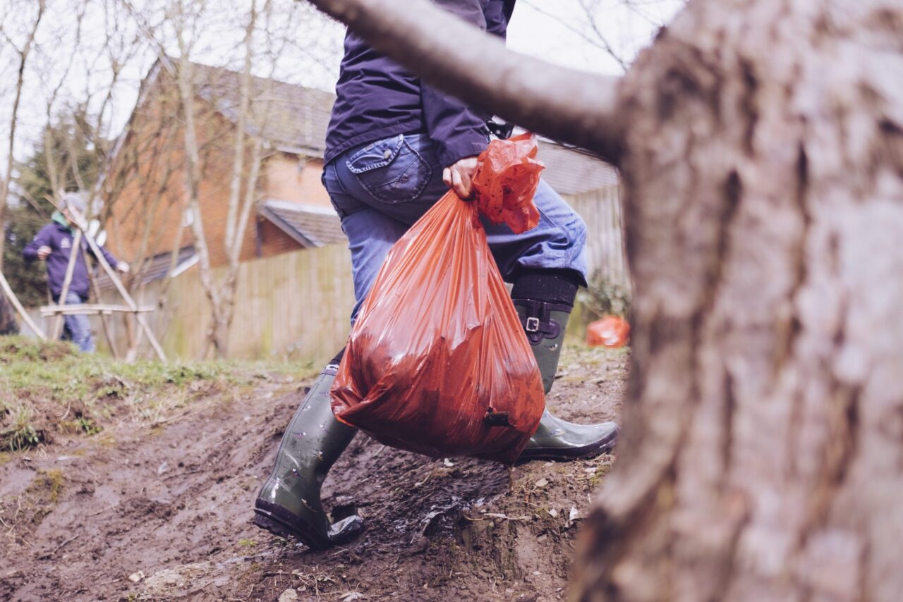 Volunteer in wellingtons carries a litter orange sack and walks behind tree