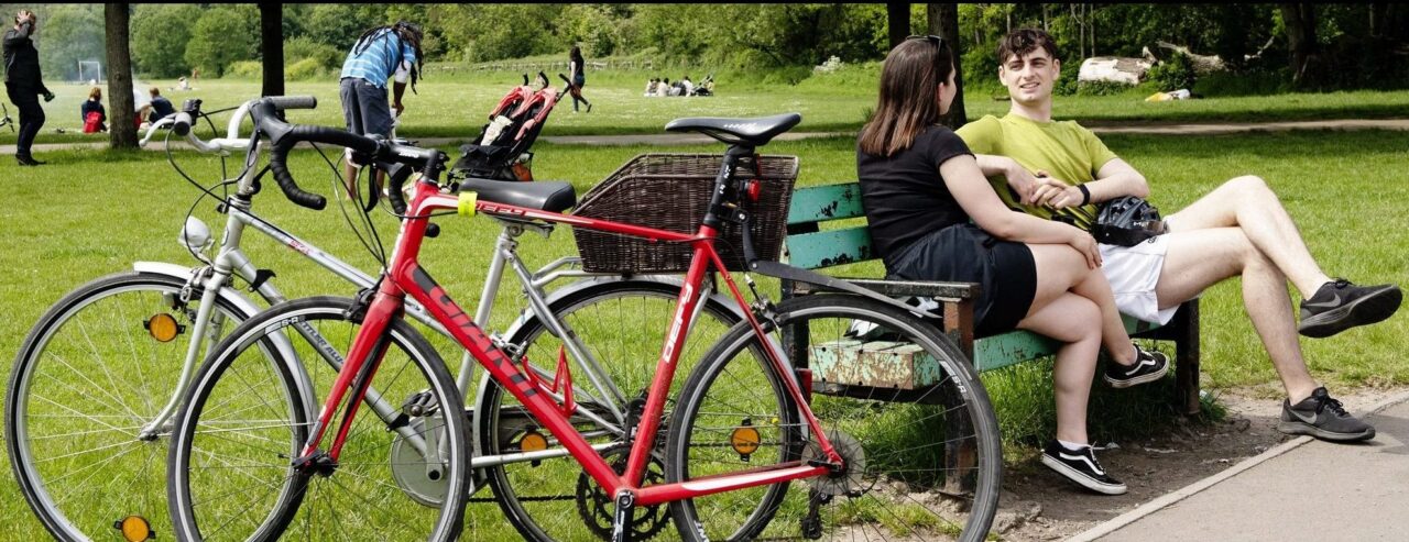 Eisteddodd dau berson ar fainc mewn parc, yn gwisgo dillad haf. Maen nhw wedi'u gosod wrth ymyl dau feic.