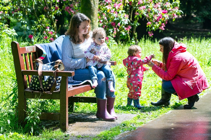 A mother and baby sat on a bench in a park. There is another mother interacting with her toddler in the background.