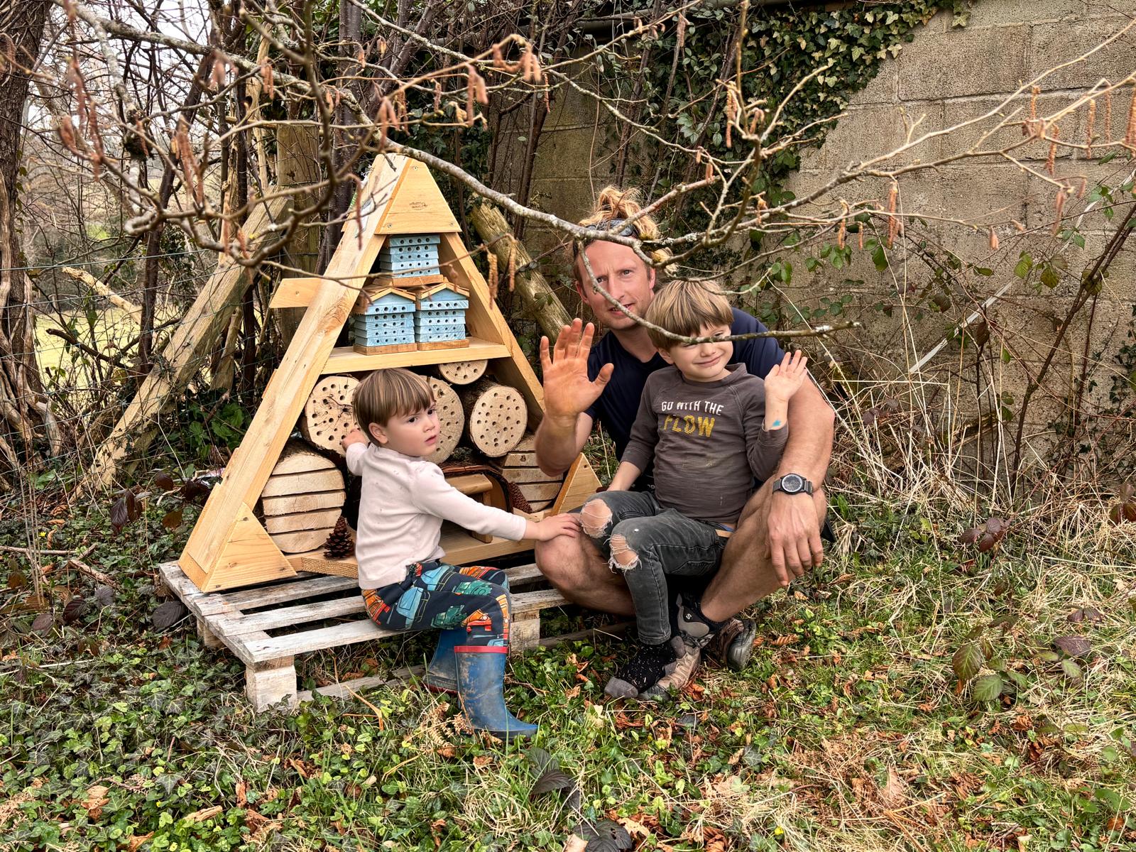 A person and two children sit in front of a wooden insect hotel, with the person waving.