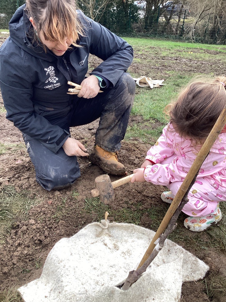A person in a dark jacket and muddy boots kneels and holds small wooden stakes while a child in a pink jacket hammers a stake into the ground next to a white fabric covering a hole.
