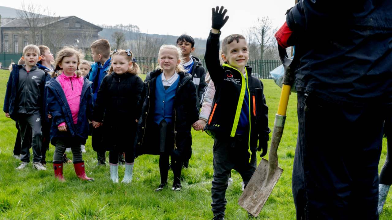A group of children in coats and school uniforms are standing in a grassy field, with one child raising a hand. An adult holding a spade can be seen on the right.
