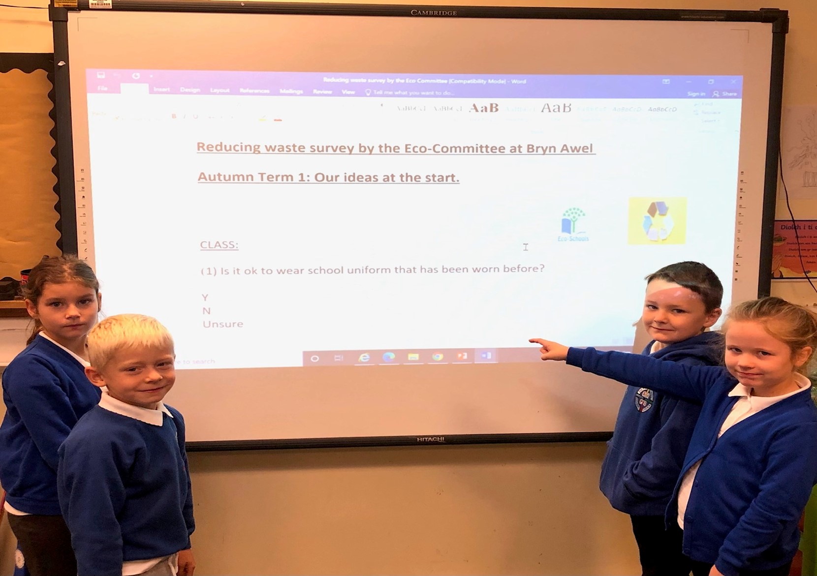 Pedwar dysgwr mewn gwisg las yn sefyll o flaen sgrin, un yn pwyntio at y sgrin.