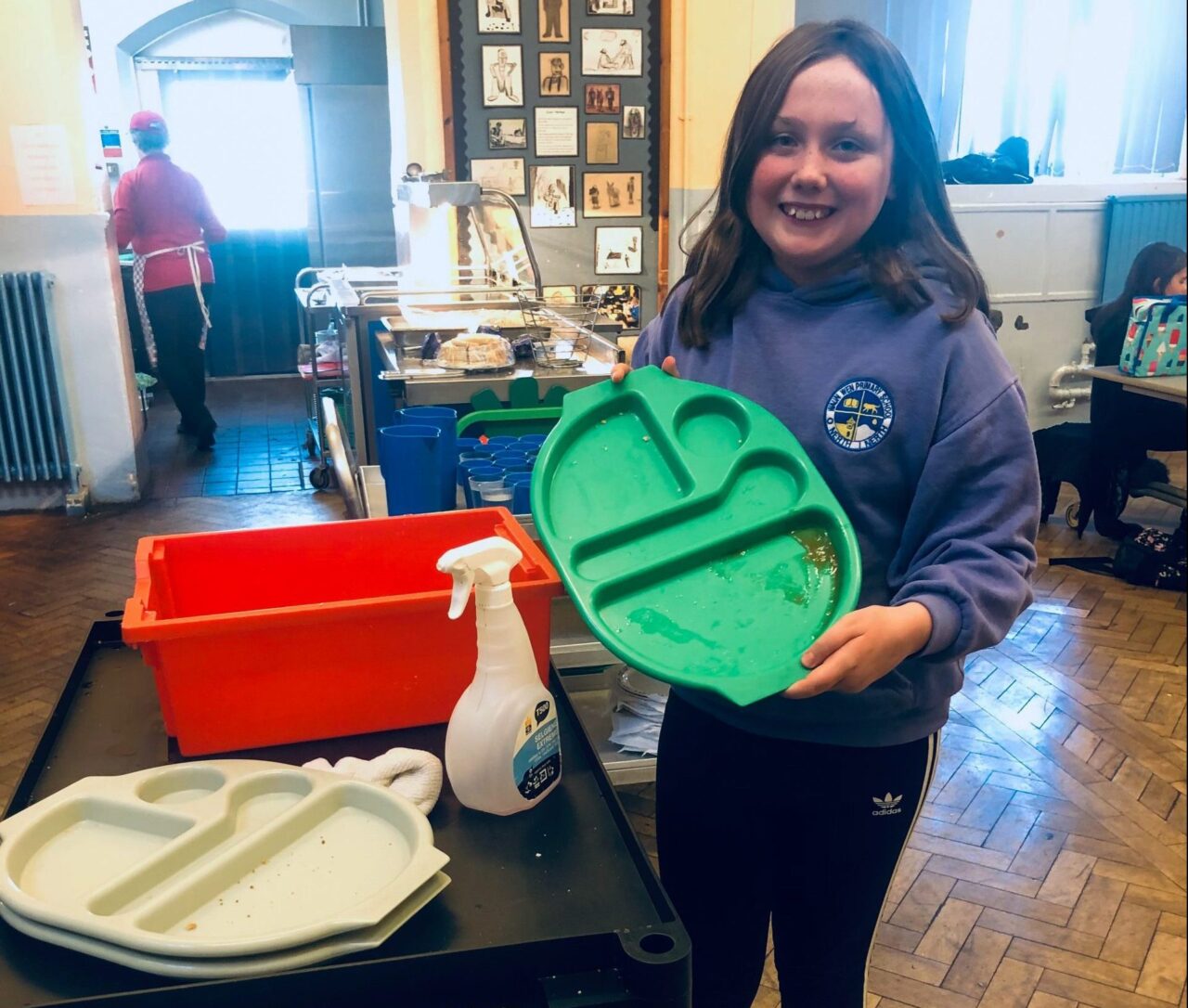 Dysgwr mewn gwisg las yn dal hambwrdd bwyd gwyrdd.