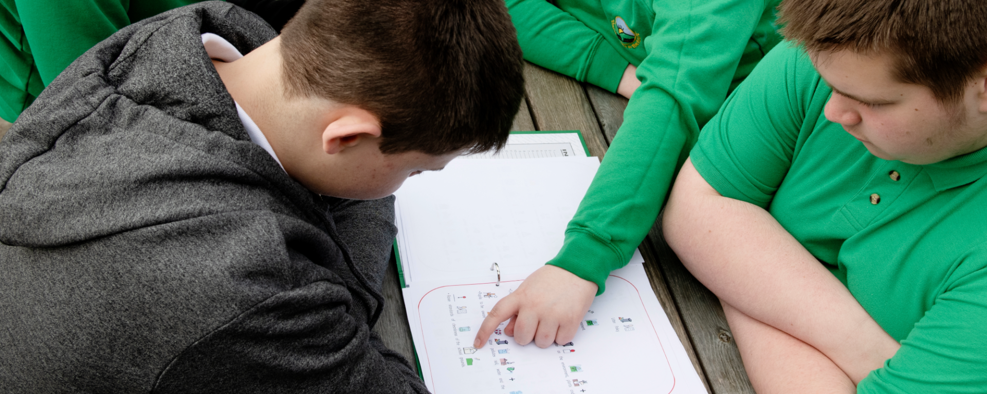 Three young people sat at a picnic bench, two of them are looking down and the other is pointing at a book.