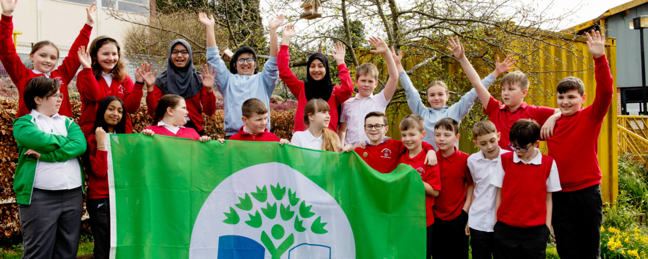 A group of young primary school children standing with their hands in the air, in front of a Eco-Schools green banner.