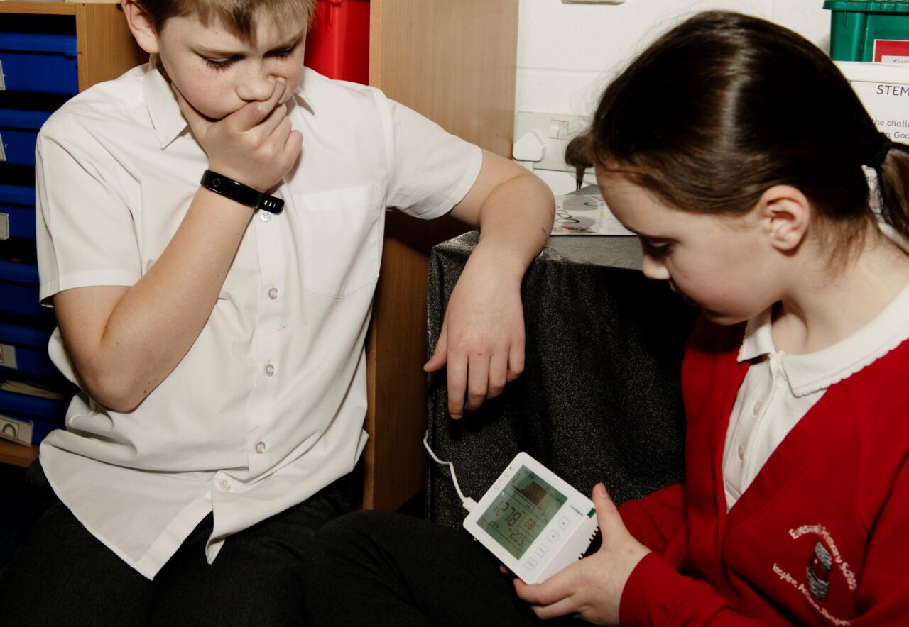 Two learners sitting in a classroom. One is holding an energy calculator wearing red uniform and the other is watching wearing a white shirt.