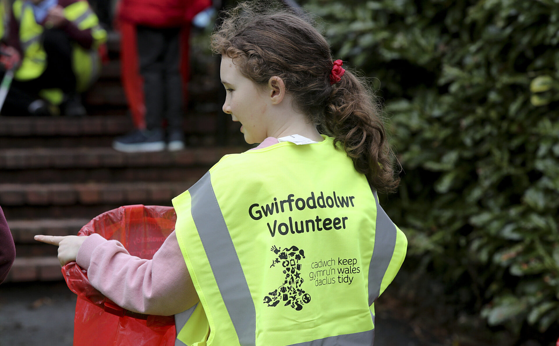 Gwirfoddolwr ifanc yn gwisgo fest gwelededd uchel yn dal bag sbwriel coch.