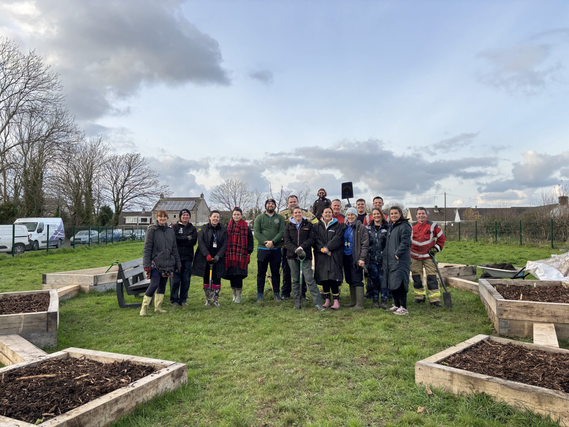 A group of people stand in a grassy area with raised garden beds in the foreground and background. A few people are holding gardening tools.