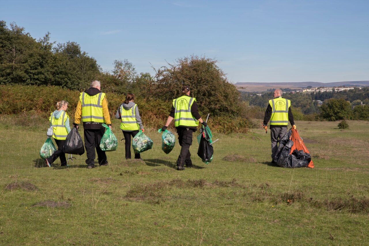 Five volunteers walking with their back to the camera, carrying litter bags and litter picks, all wearing hi-vis vests.