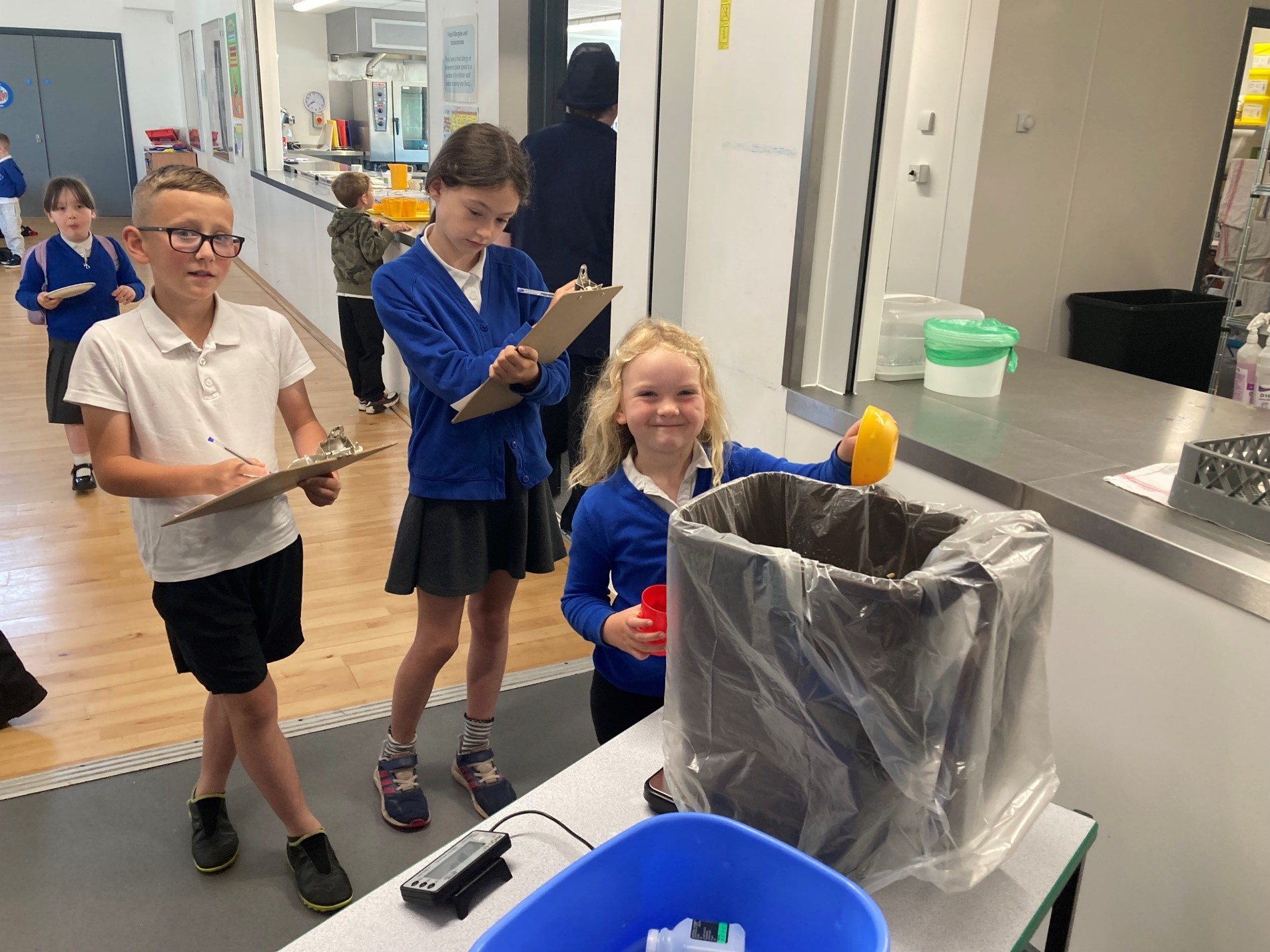 Dau ddysgwr yn dal clipfwrdd yn sefyll y tu ôl i ddysgwr ifanc yn gwenu ac yn dal powlen wrth ymyl bin du. Maen nhw'n gwisgo gwisg ysgol las a gwyn.