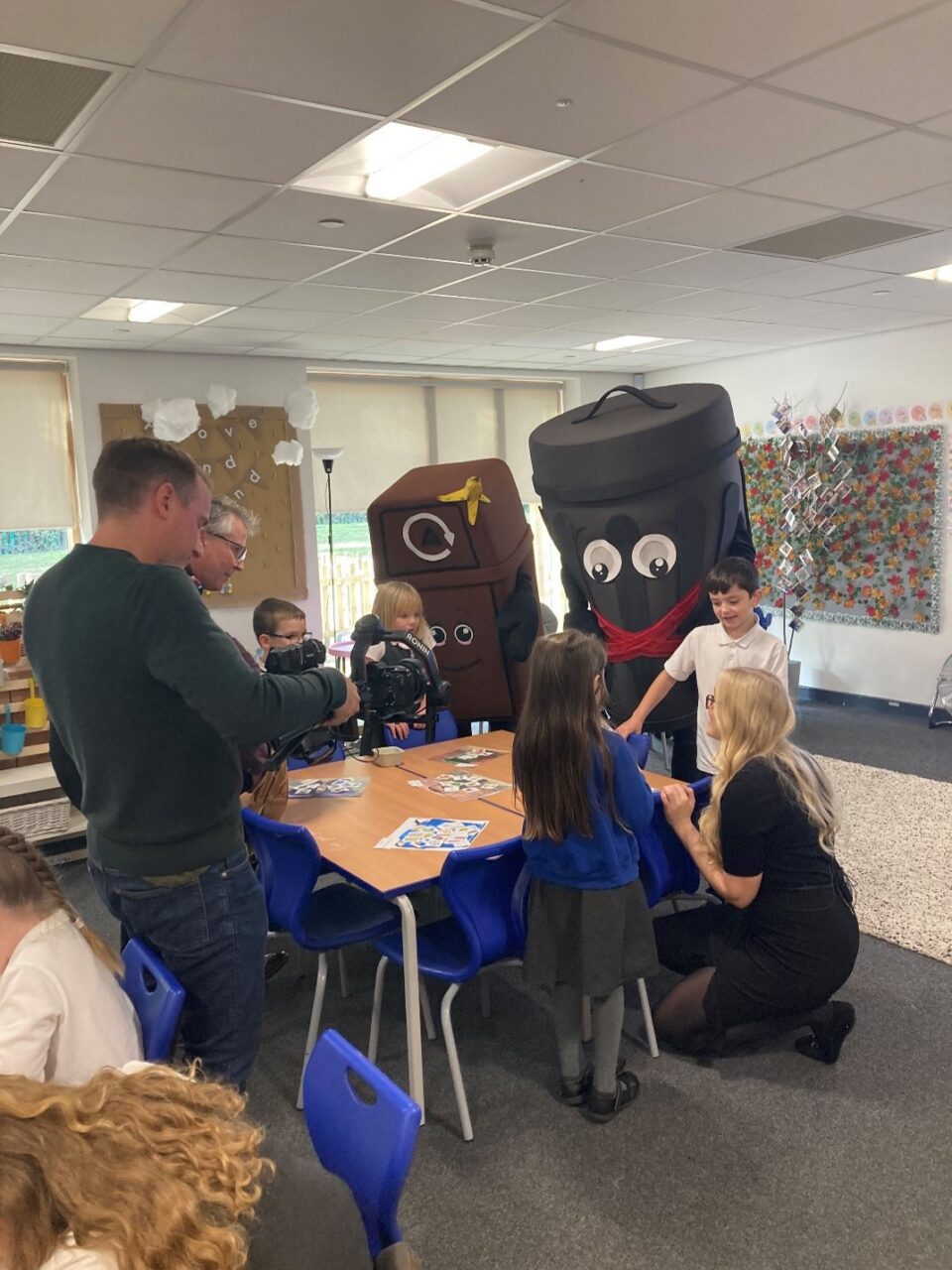 Roedd pobl ifanc yn eistedd wrth fwrdd, ac mae camera yn sefyll o'u blaenau. Y tu ôl i'r bobl ifanc mae gwisgoedd bin brown a du.