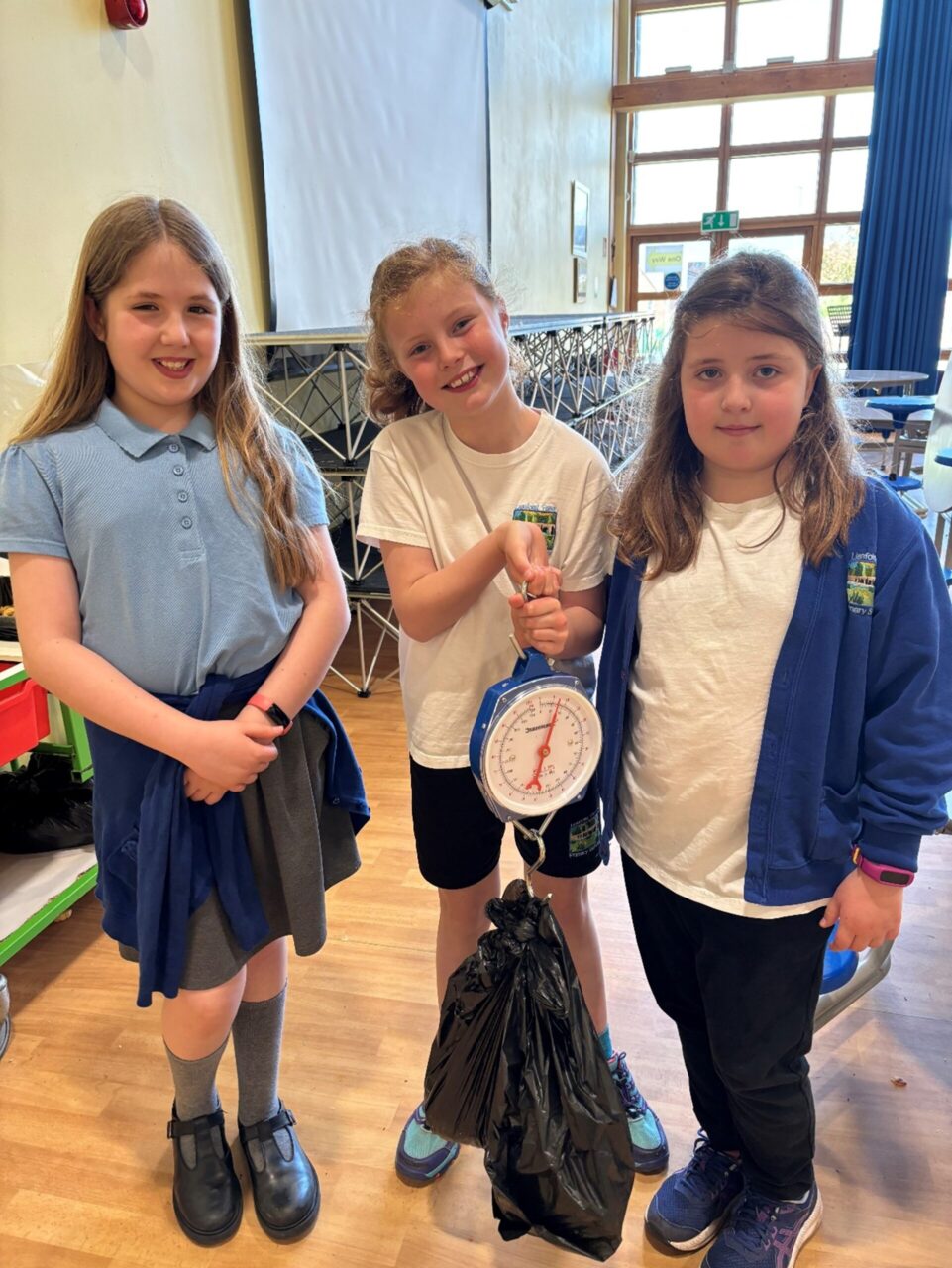 Three young learners wearing blue school uniform. One of them is holding scales with a black bag at the end of it.