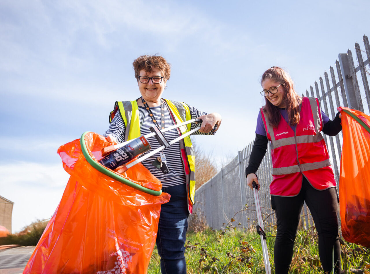 Dwy wirfoddolwr benywaidd yn gwenu, yn defnyddio codwyr sbwriel i roi sbwriel mewn bagiau sbwriel coch