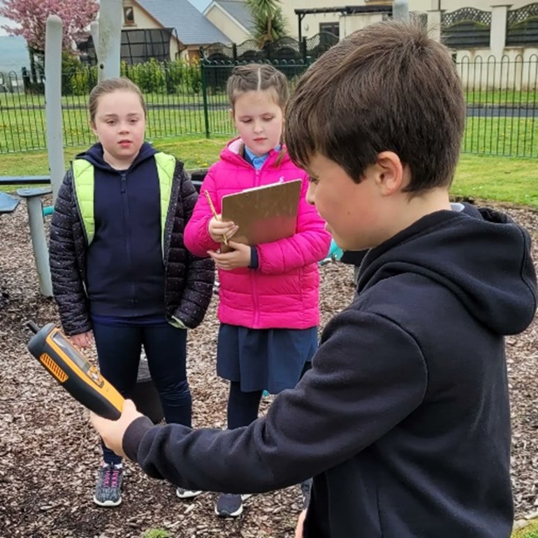 Mae plentyn yn dal dyfais felen tra bod dau blentyn arall yn arsylwi, un yn dal clipfwrdd.