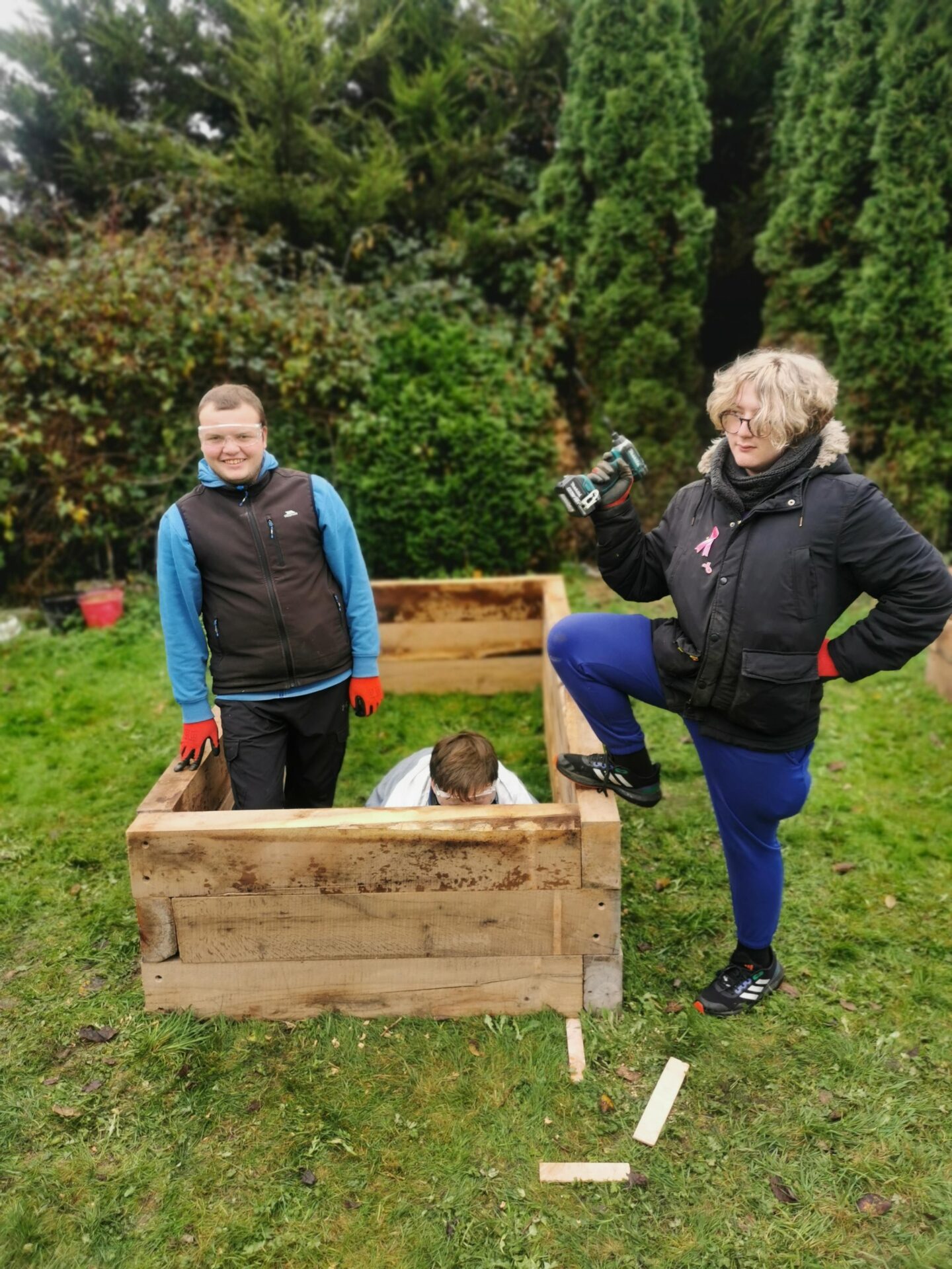 Two people are inside a raised bed wearing protective goggles, one of them is standing and the other is crouching. A third person is standing beside the raised bed holding a drill wearing a coat.