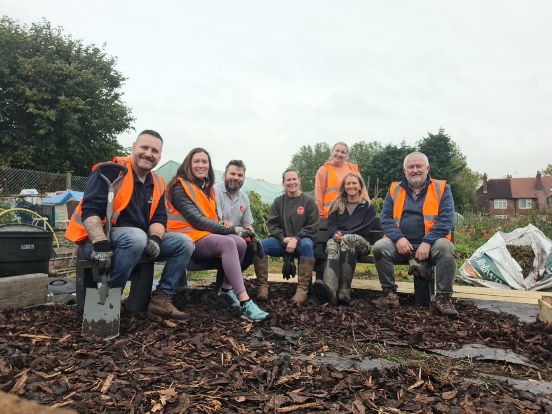 Grŵp o bobl yn gwenu yn eistedd i lawr mewn gardd, yn gwisgo siacedi oren llachar.