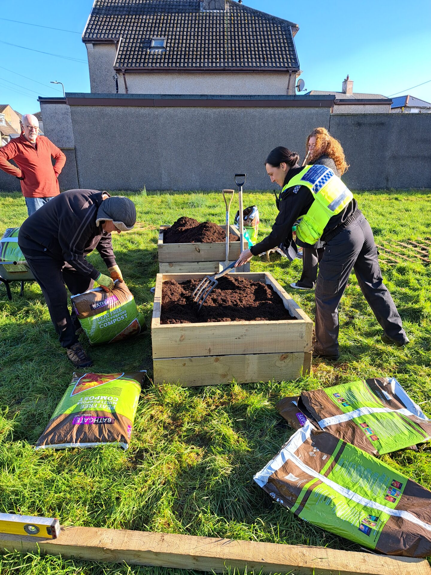 Dau berson wrth wely uchel, mae un ohonyn nhw'n gwisgo lifrai heddlu yn dal fforch gardd. Mae'r llall yn agor bag o gompost.