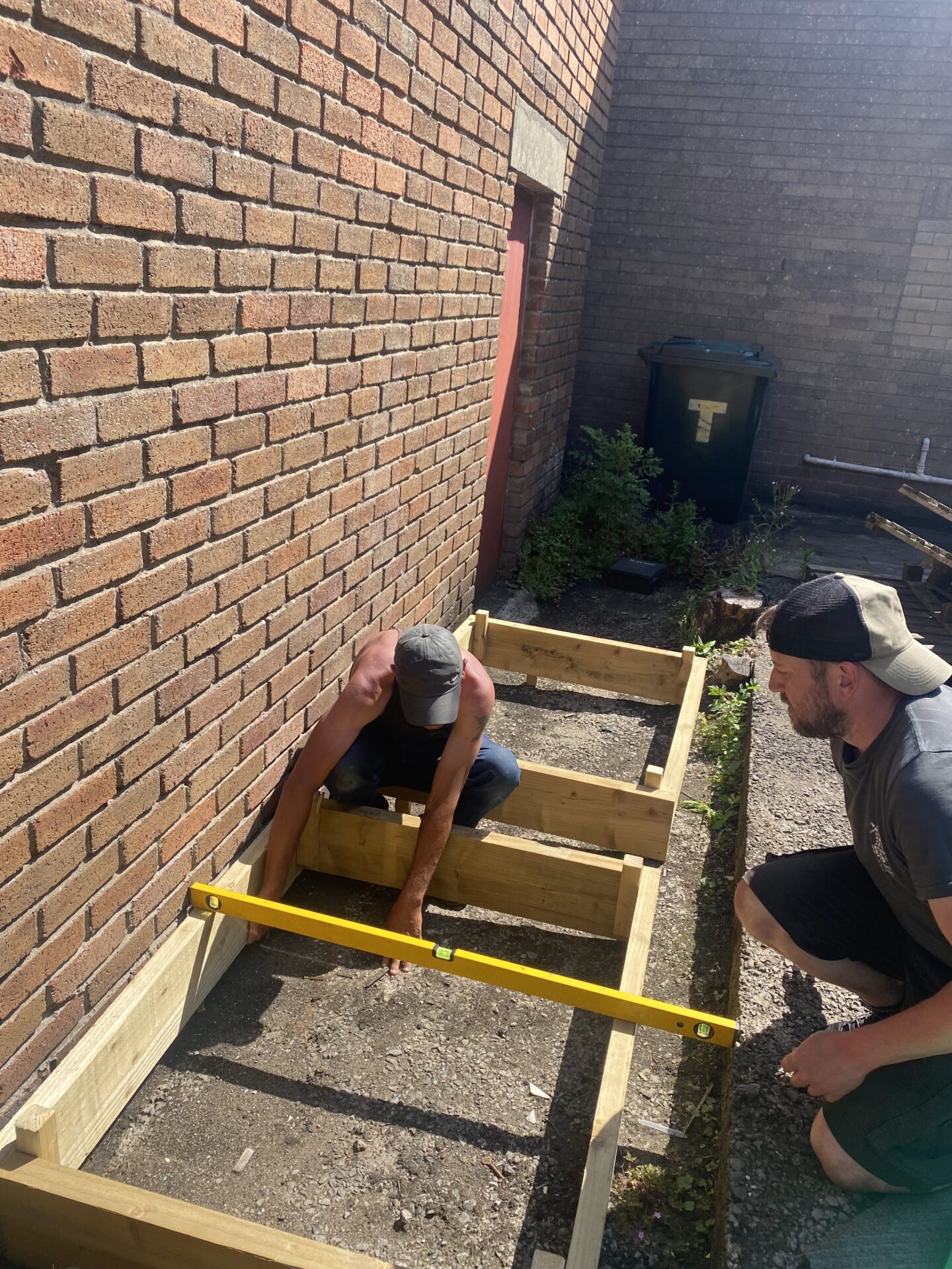 Dau berson yn dal planciau pren, maen nhw ill dau yn gwisgo capiau pêl fas y tu allan i adeilad brics.