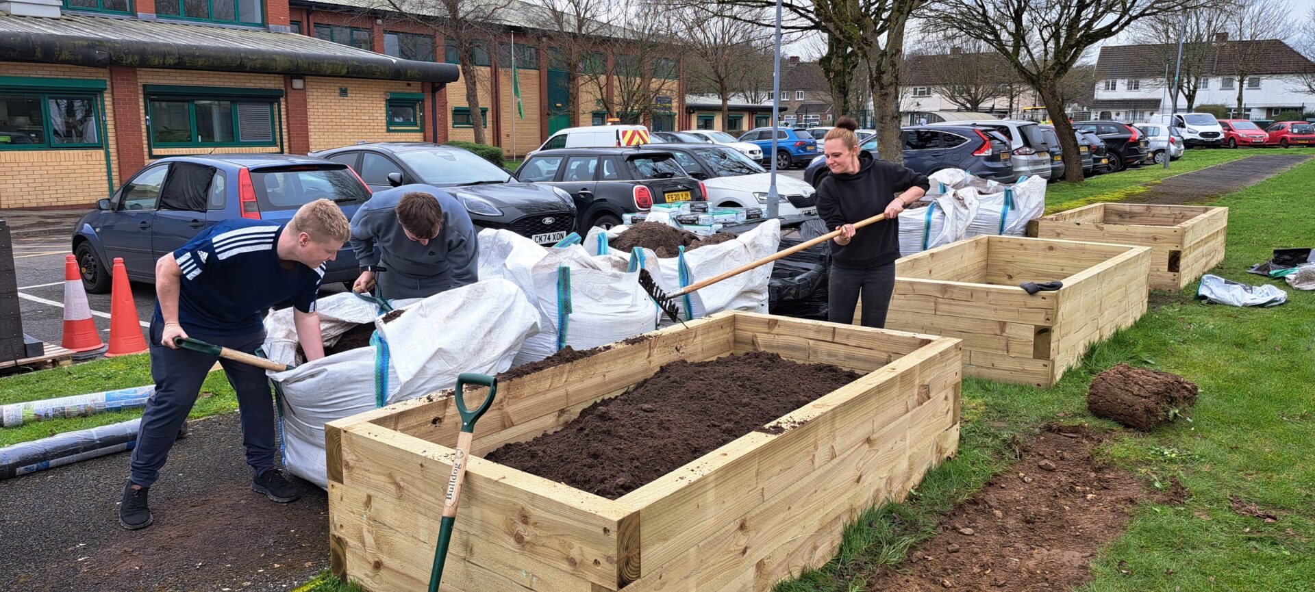 Three people by a raised bed, one of them is holding a garden fork and the other two are digging in a bag of compost.