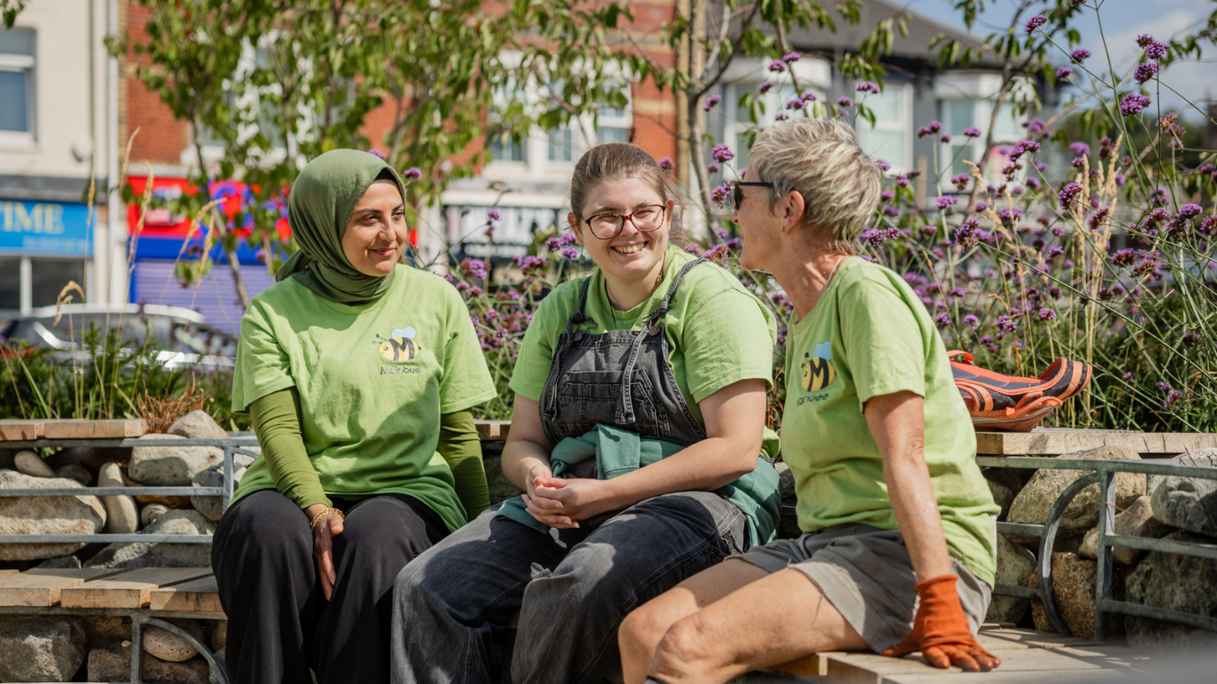 Three individuals in green shirts are sitting on a stone bench and talking. One is wearing a green hijab, another is wearing glasses and dark overalls, and the third is wearing glasses and orange gloves.