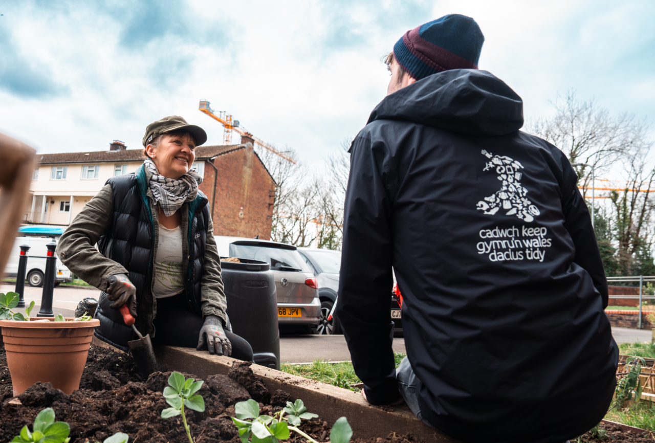 A person in a cap and vest gardening while another person in a black jacket with text and graphics on the back looks away.