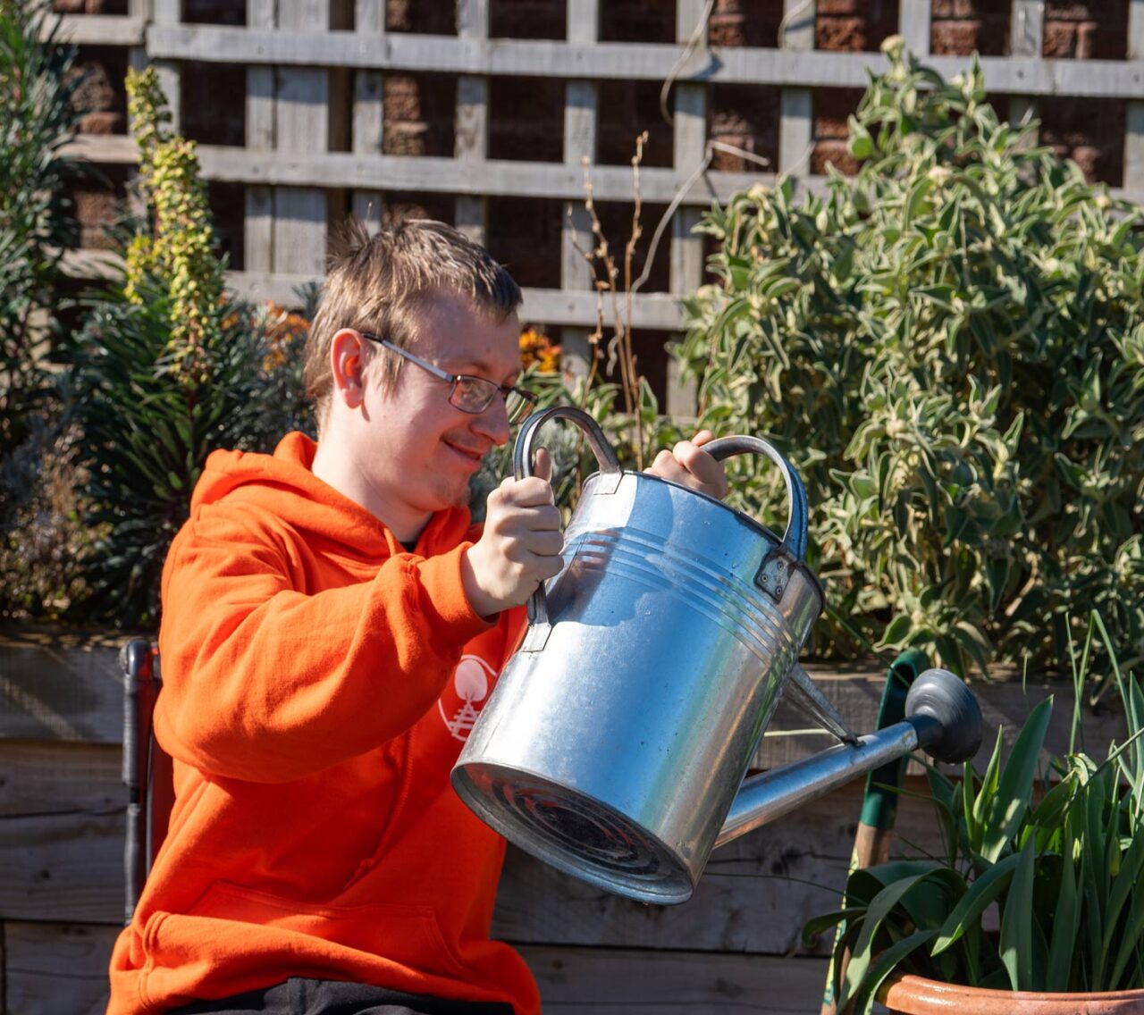 A person wearing an orange hoodie waters plants in a garden with a metal watering can.