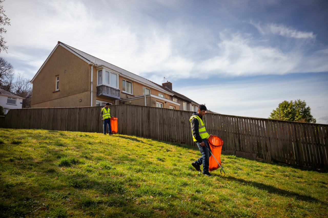 Two young volunteers in hi-vis vest, holding litter pickers and red bags, walking down a grassy slop on a housing estate.