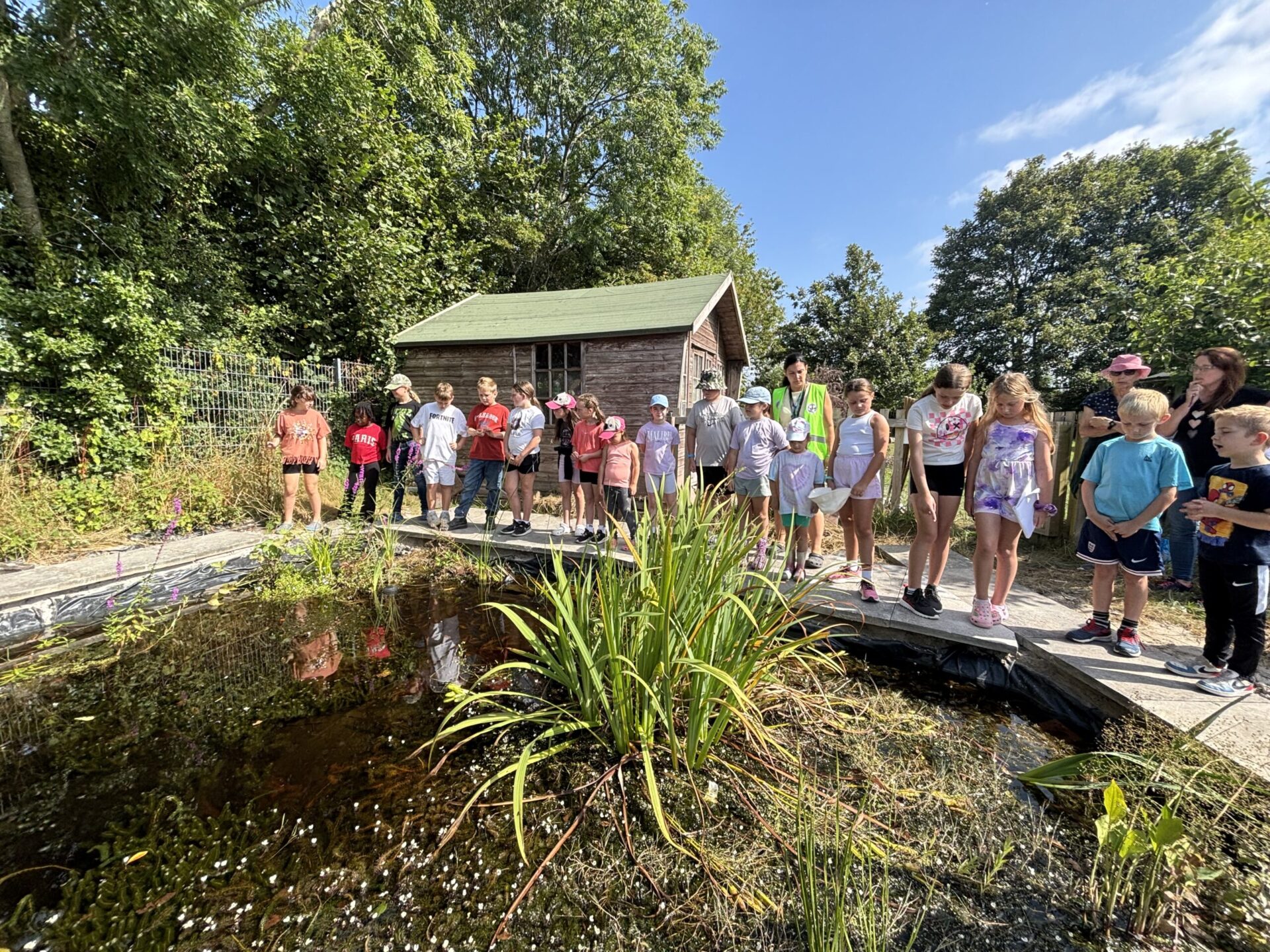 A group of young learners lined up by a pond, in front of a shed with blue skies.