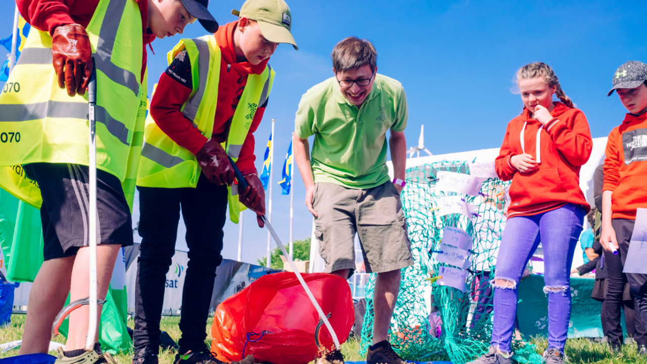 Several individuals, some wearing safety vests and gloves, are gathered around a large red bag on the ground, holding litter picking kit.