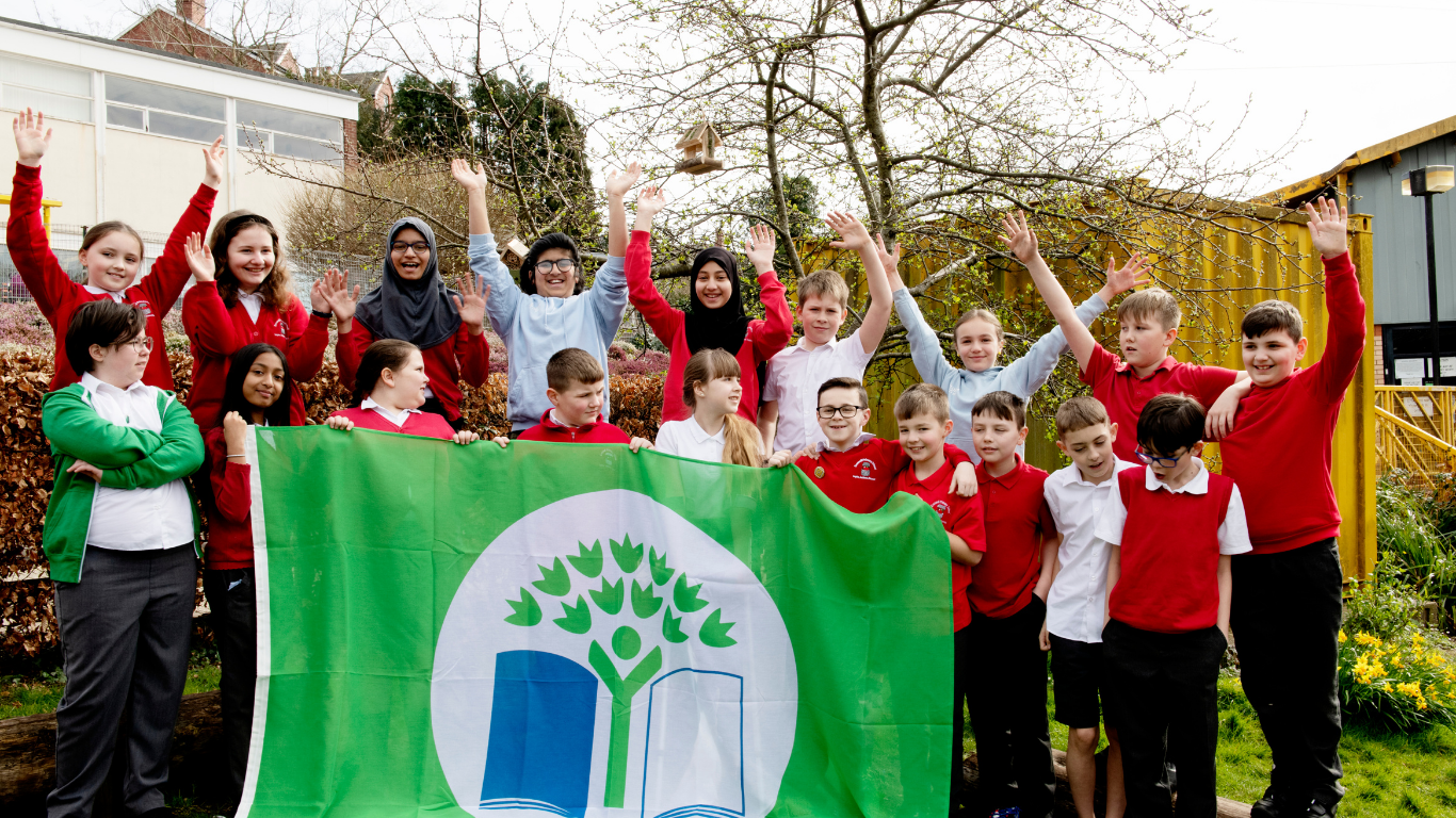 A group of children in school uniforms hold up a large green flag with the Eco-Schools logo.