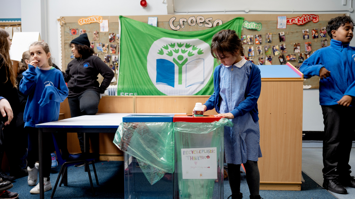 A group of children in a classroom are participating in a recycling activity. One child in a blue gingham dress is placing a small item into a red recycling bin, while other children watch. A large green banner with the Eco-Schools logo behind them.