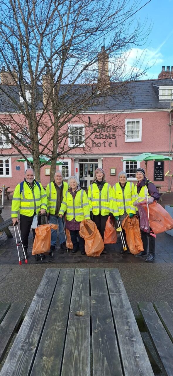 A group of six volunteers, outside a red building, all smiling carrying red litter bags