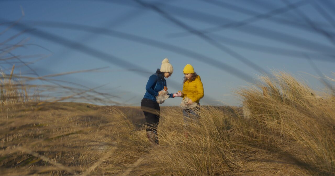A mother and teenage daughter stand together in sand dunes inspecting litter they've collected