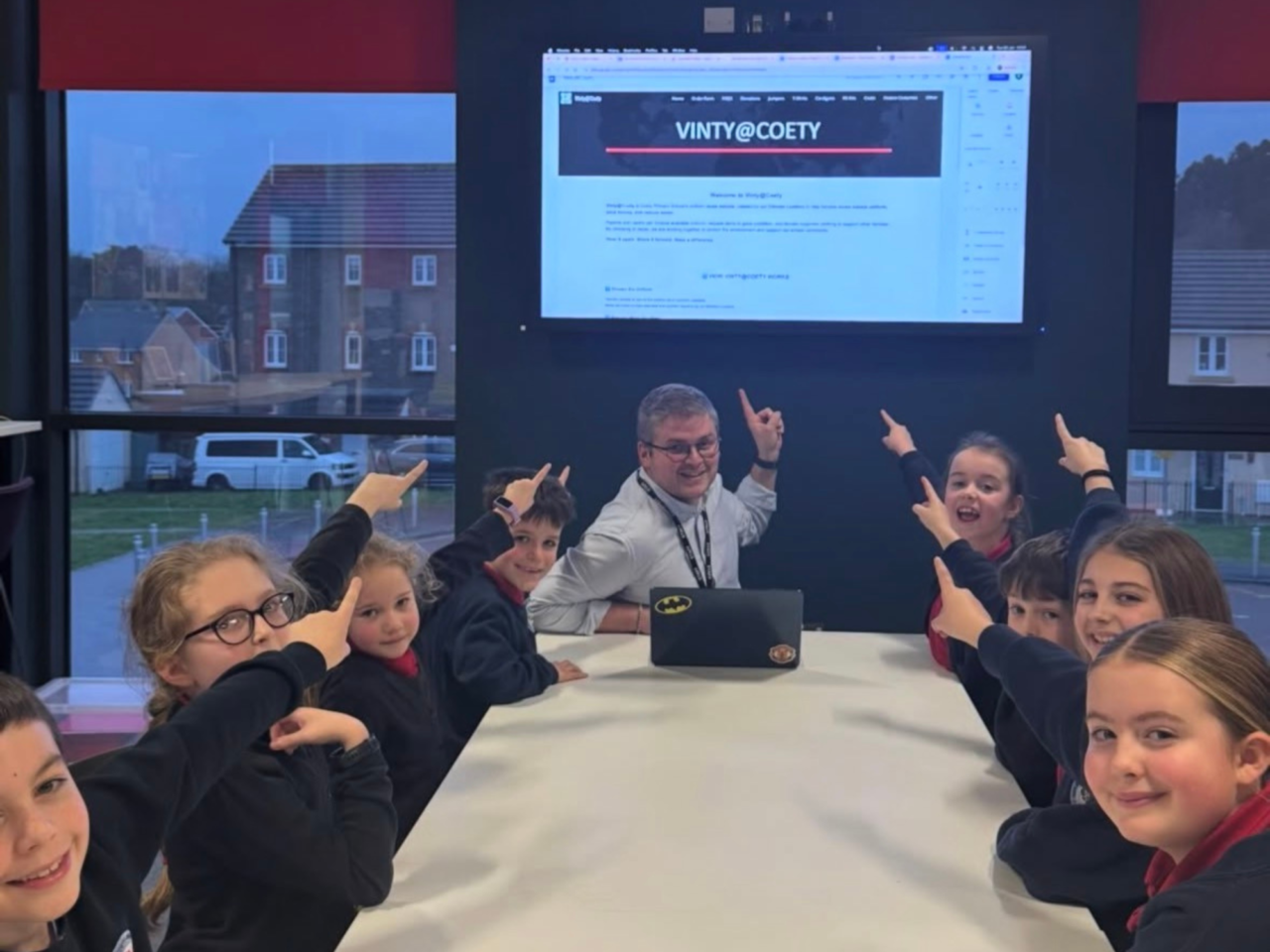 A man and several children are pointing at a large screen displaying "VINTY@COETY".