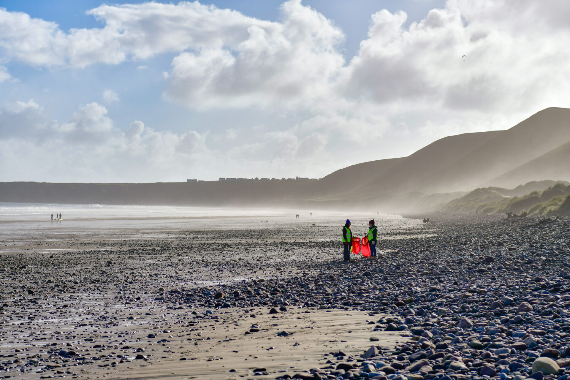 Dau unigolyn yn siarad tra’n codi sbwriel ar draeth yng Nghymru ar ddiwrnod braf