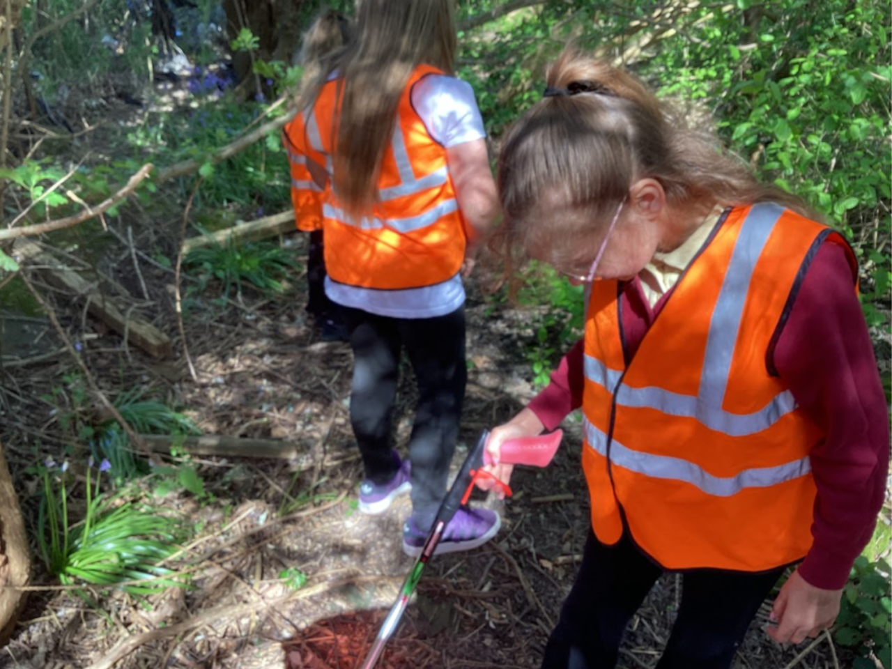 Two young people wearing orange hi vis vests, one of them has their back turned to the camera and the other is holding a litter grabber.
